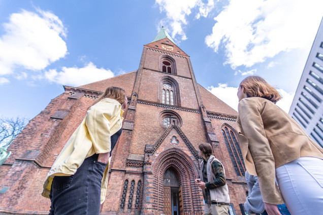Gruppe aus vier Erwachsenen in einer Stadtführung in Kiel, Kirche aus Backstein im Hintergrund, Froschperspektive